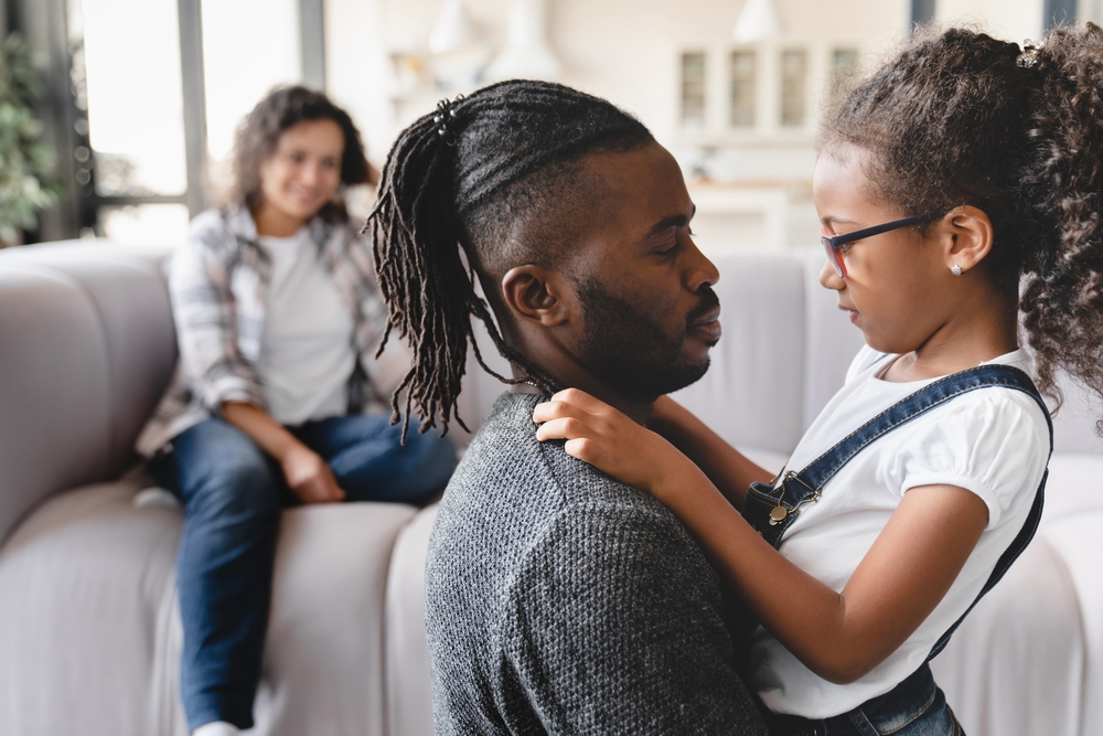 Loving african-american parents couple having fun with small preteen daughter, spending time together at home, hugging and embracing. Family bonding.