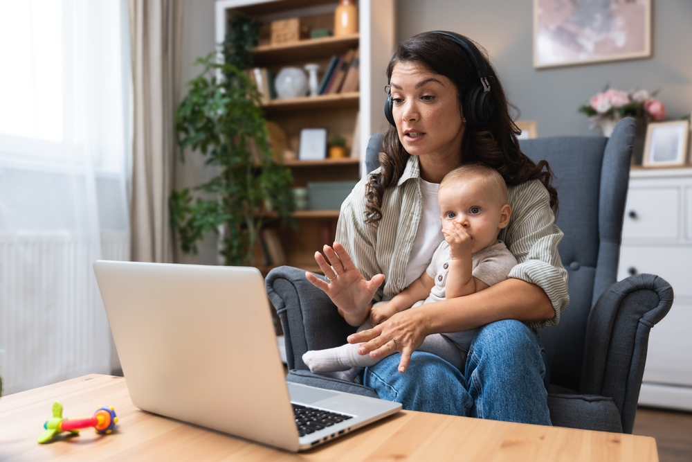 Single mother with baby on lap speaks emotionally via laptop video call to ex-husband about overdue child support, alimony, sitting in armchair wearing wireless headphones, facing financial stress.