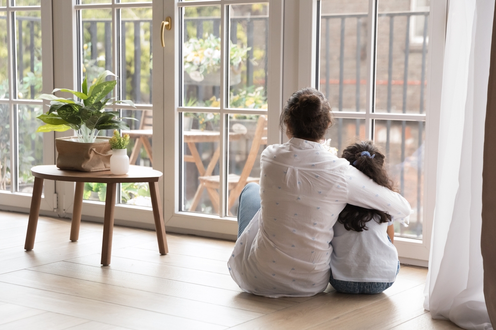 Back view of black haired young mom and little daughter girl sitting on floor at home, hugging with love, care, looking at panoramic window, cozy balcony. Motherhood, real estate concept