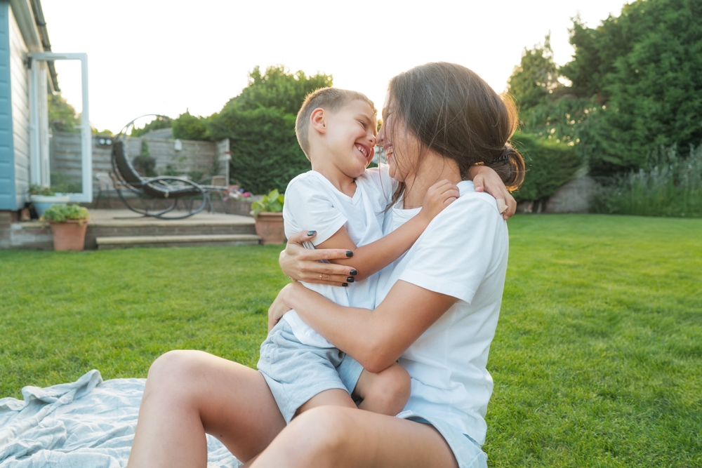 Cheerful cute preschooler boy kid having fun with his mum, embracing, laughing on the green grass.