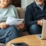 A mature couple, mid-50s to early 60s, sitting separately in a quiet, modern living room with neutral tones. Each person has financial documents or a laptop in front of them, suggesting retirement planning and legal consultation.