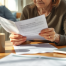 A middle-aged woman sits at a kitchen table, holding legal documents. An elderly parent rests in the background.