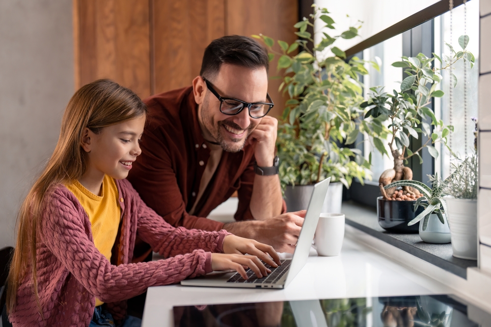Handsome father wearing glasses helping his elementary age daughter with her homework while she's sitting at the desk at home and typing on a laptop .