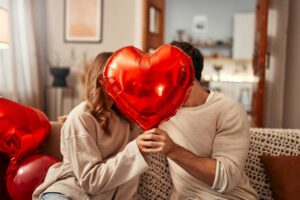 Happy Valentine's Day. Young couple in love holding a heart-shaped balloon, covering themselves with it while kissing, sitting on the sofa in the living room at home. Romantic evening together.