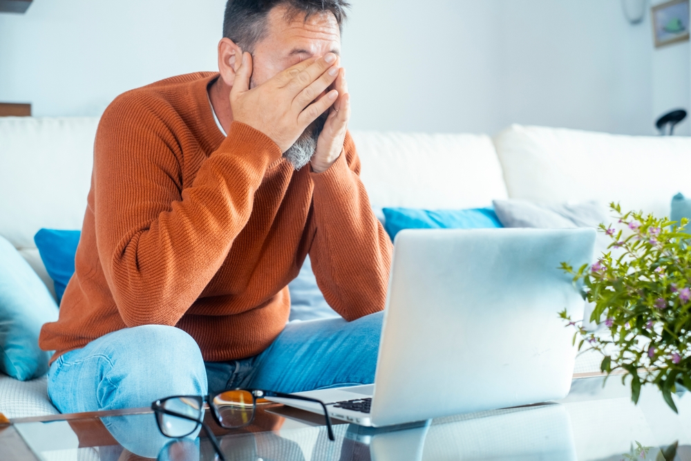 Exhausted man sitting on the sofa at home, reading troubling emails from his lawyer about divorce and family bankruptcy, feeling overwhelmed by financial problems and the stress online notifications