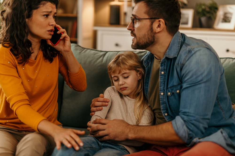 Sad little girl sitting between parents on sofa while her mother talking on phone with social worker about custody and child support after divorce