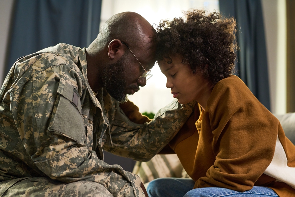 Black middle aged man in military uniform comforting Black young adult woman at home, both sitting close with foreheads touching, showing emotional family support and connection